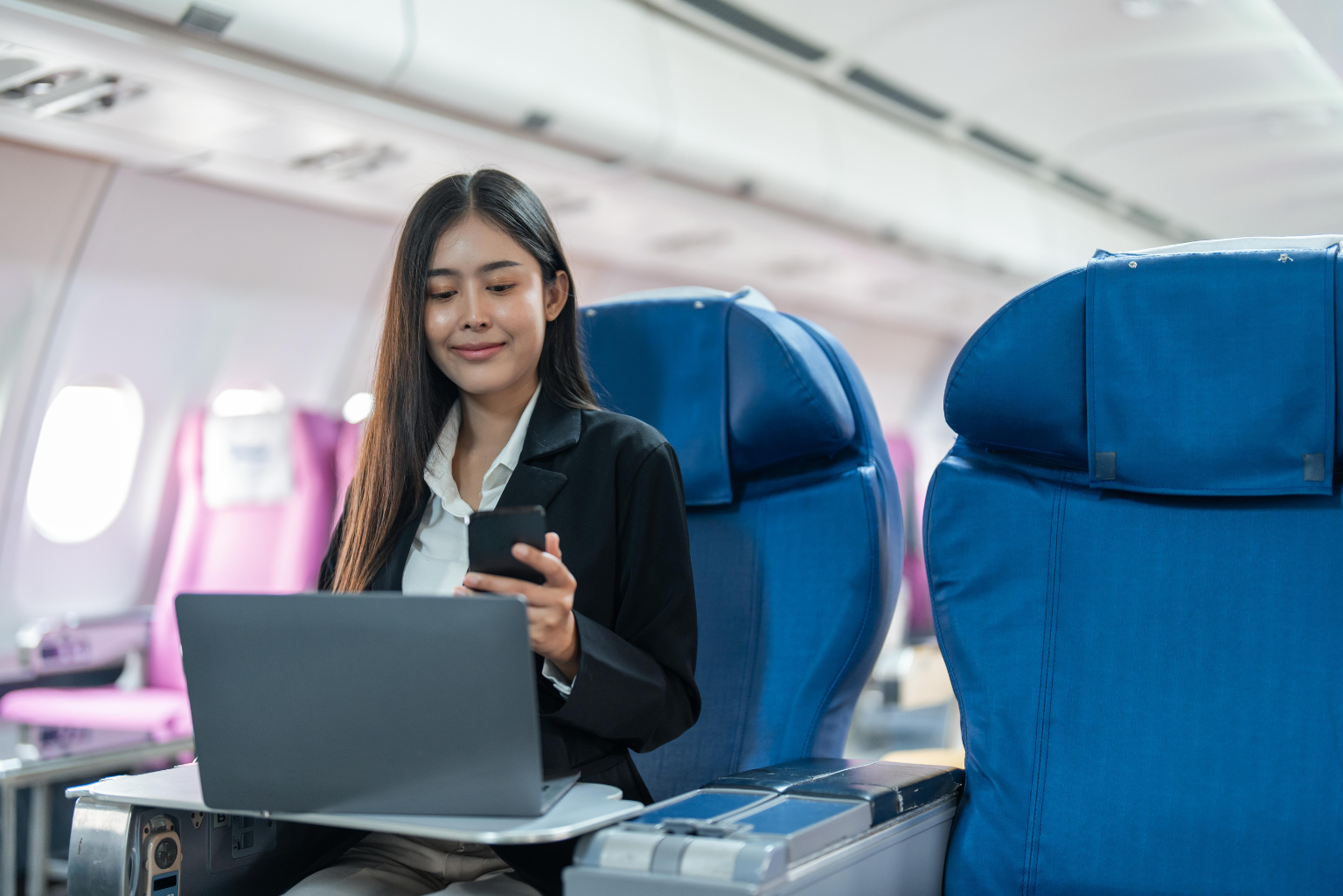 A young woman in business casual attire on an airplane working from her phone and her laptop.