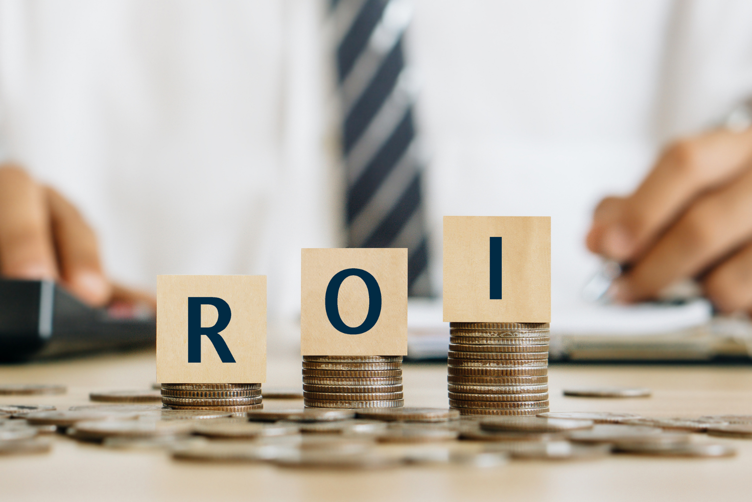 R-O-I written on wooden blocks and stacked on top of increasingly larger piles of coins, all on top of a desk covered in scattered coins where a businessperson sits.