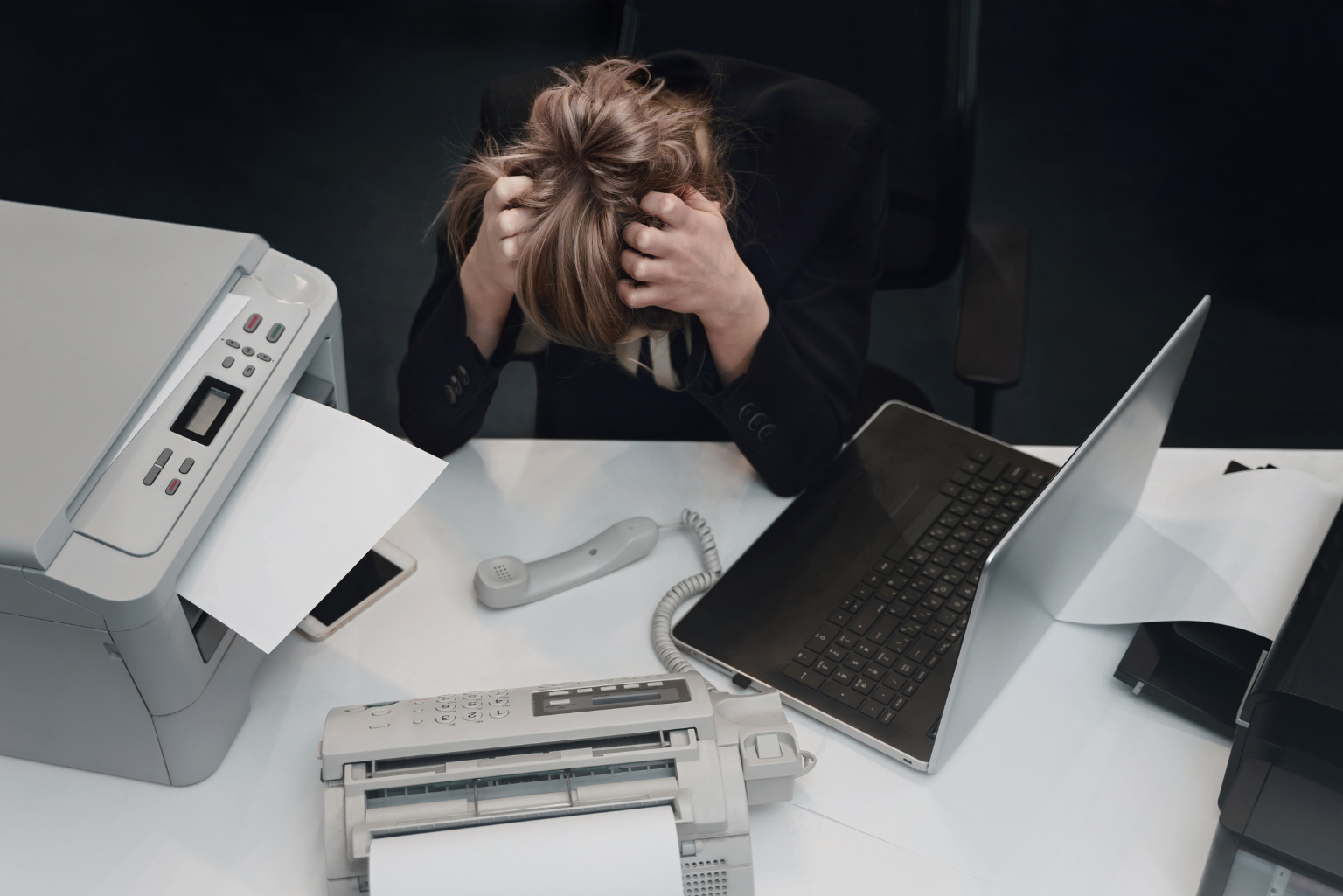 A stressed office employee at a desk surrounded by legacy technology: a fax machine, a large printer, an old laptop.