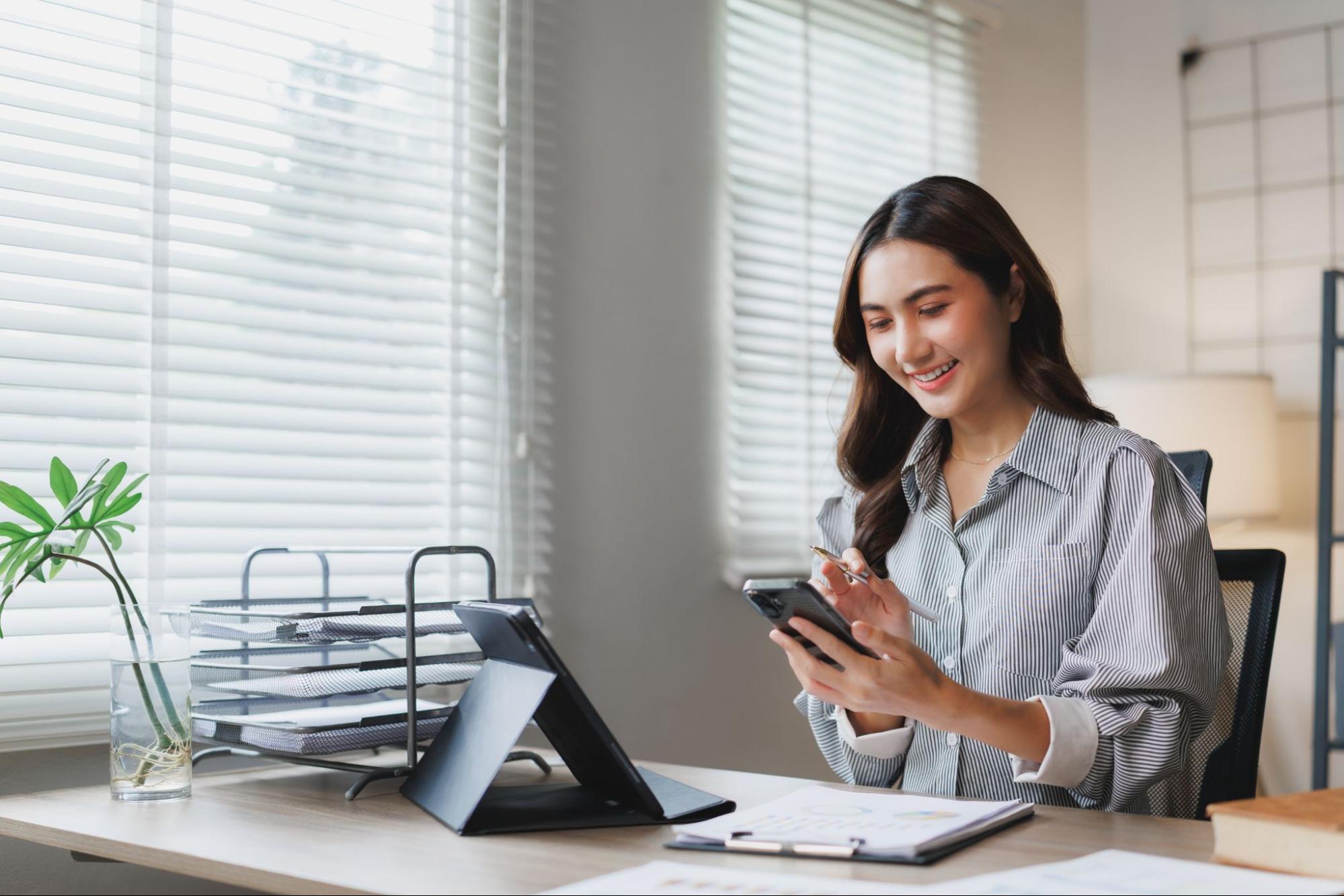 Smiling businesswoman at a desk in her office using a smartphone and tablet for work.